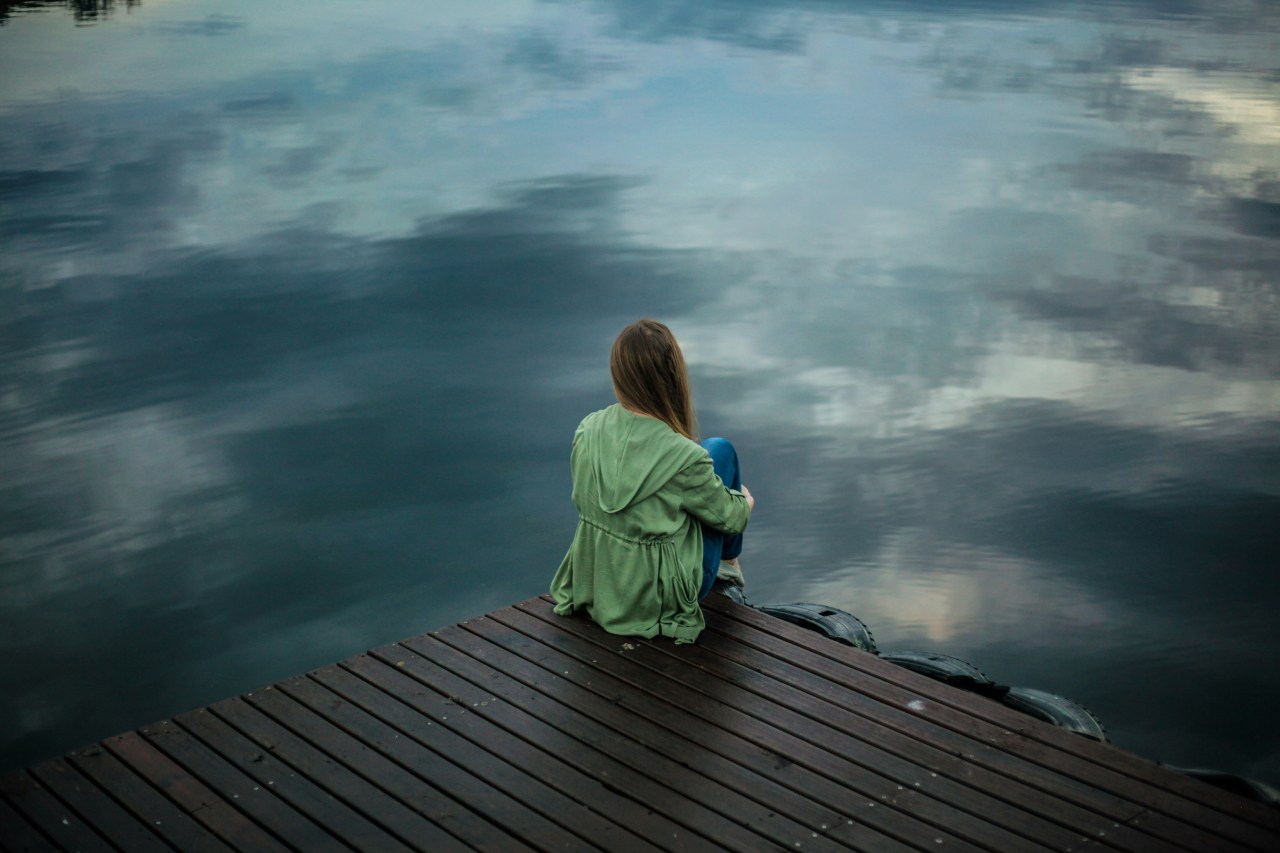 A woman sits alone at the edge of a wooden dock, knees drawn to her chest, gazing out over still water that mirrors a clouded sky.