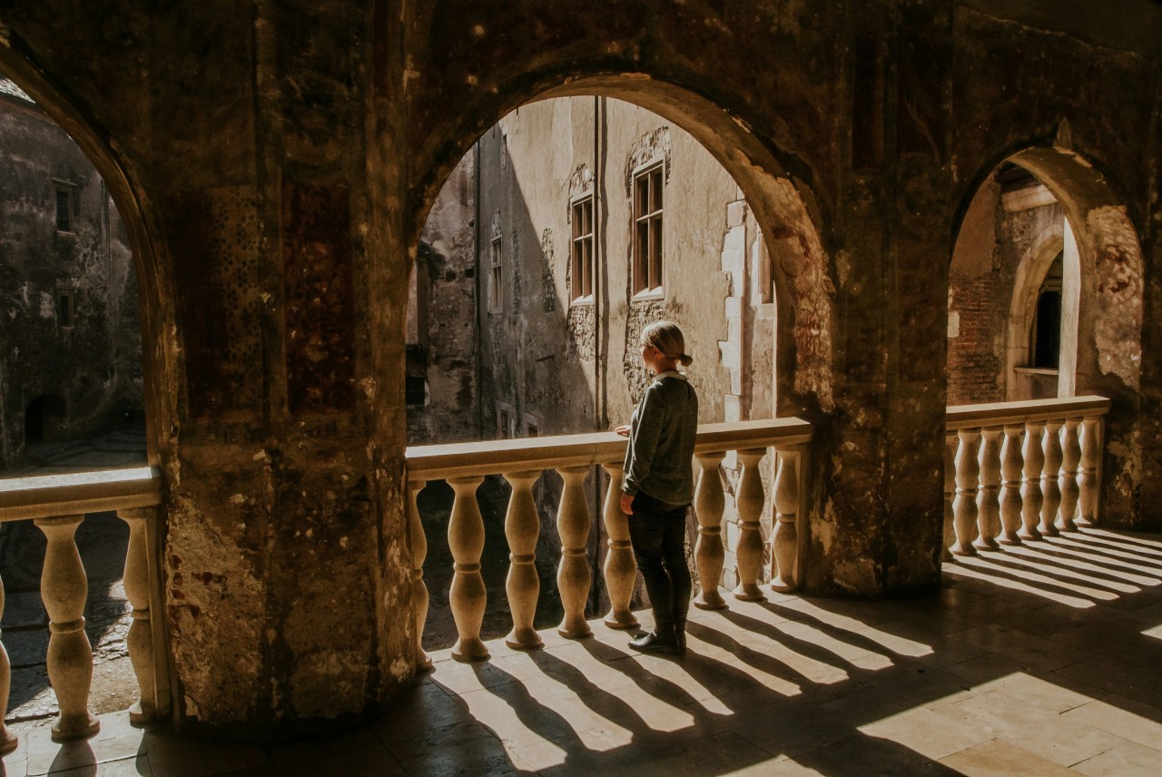 A woman stands alone at a weathered stone balustrade, looking out through an arched colonnade toward an old sunlit courtyard.